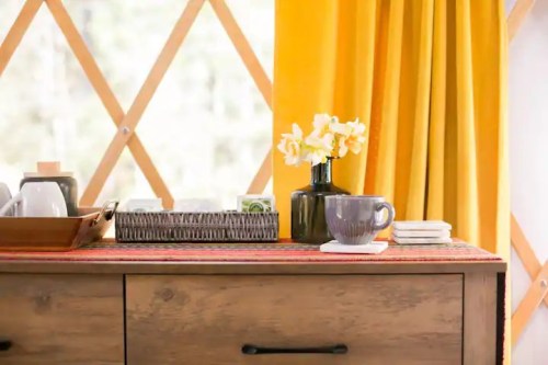 A cozy interior scene featuring a wooden dresser, a vase of flowers, a mug, and yellow curtains.