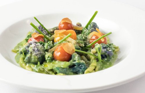 A plate of green pasta with cherry tomatoes, garlic chips, and herbs, garnished with grated cheese.