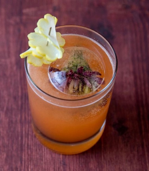 A refreshing cocktail with a floral garnish and a large ice cube, served in a clear glass on a wooden surface.