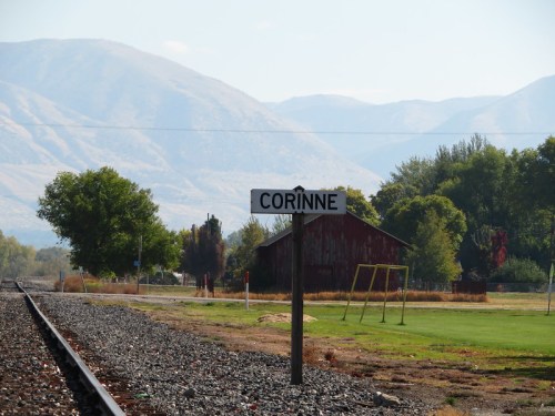 Sign for Corinne near railroad tracks, with mountains and a barn in the background on a clear day.