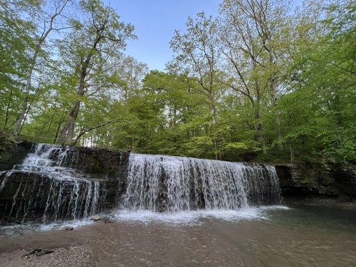 A serene waterfall cascading over rocks, surrounded by lush green trees and a clear blue sky.