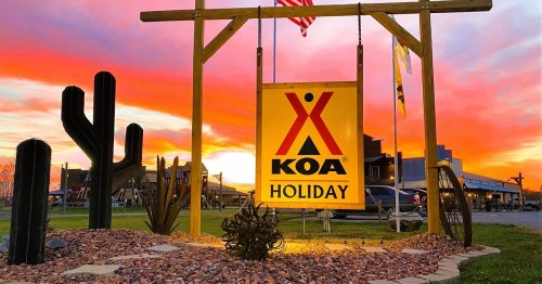 A KOA Holiday sign illuminated at sunset, with cacti and a colorful sky in the background.