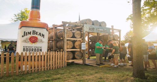 A large Jim Beam bottle stands beside a display of bourbon barrels and a crowd at an outdoor event.