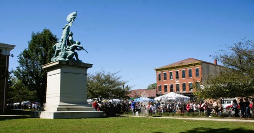 A statue of a soldier on a pedestal with a crowd in the background, enjoying a sunny day at an outdoor event.