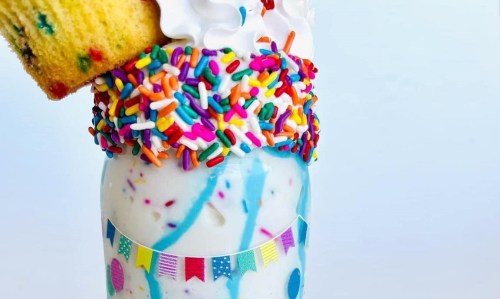 A colorful milkshake topped with whipped cream, sprinkles, and a piece of cake, served in a jar with festive decorations.