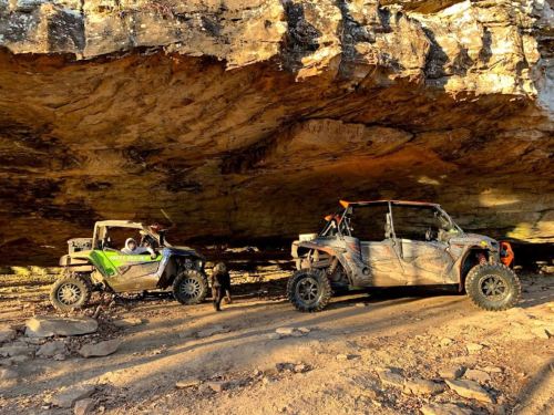 Two off-road vehicles parked under a rocky overhang, with a dog nearby on a dirt path.