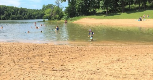 A sandy beach by a calm lake with people swimming and enjoying the sun on a clear day.