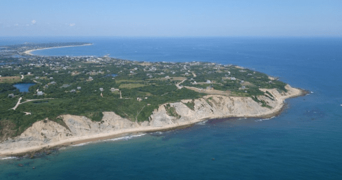 Aerial view of a coastal landscape with cliffs, sandy beaches, and lush greenery under a clear blue sky.