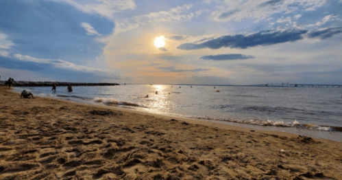 A serene beach scene at sunset, with gentle waves, sandy shore, and people enjoying the water under a cloudy sky.