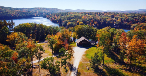 Aerial view of a lakeside cabin surrounded by colorful autumn foliage and a serene body of water.