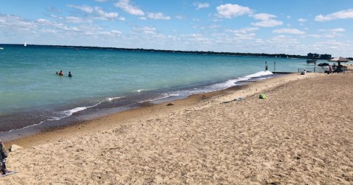 A sandy beach with calm blue water, a few people swimming, and a dock extending into the sea under a partly cloudy sky.