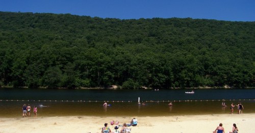 A sandy beach by a calm river, with people swimming and relaxing under a clear blue sky and green hills in the background.