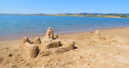 A sandy beach with a detailed sandcastle beside calm water and distant hills under a clear blue sky.