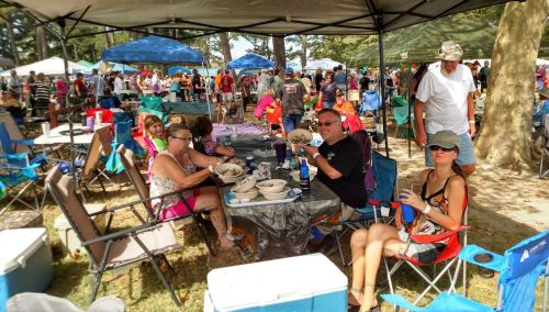 A group of people enjoying food at a crowded outdoor event under tents, with many attendees in the background.
