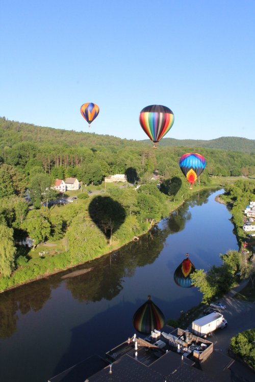 Colorful hot air balloons float over a serene river, casting shadows on the water and surrounding greenery.