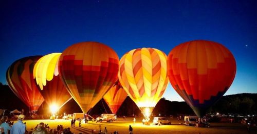 Colorful hot air balloons illuminated at night, set against a dark blue sky with a crowd watching below.