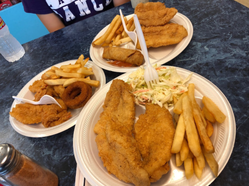 Three plates of fried food: fish fillets, fries, onion rings, and coleslaw, with a drink in the background.