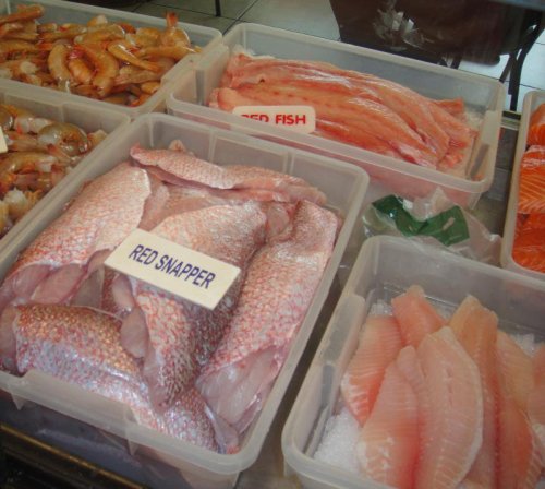 Display of fresh seafood in containers, including red snapper and various shrimp. Labels indicate types of fish.