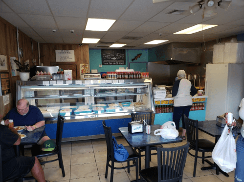 A casual diner interior with tables, a counter, and a display case filled with food. Customers are seated and ordering.