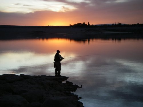 A silhouette of a fisherman standing by the water at sunset, with vibrant colors reflecting on the surface.