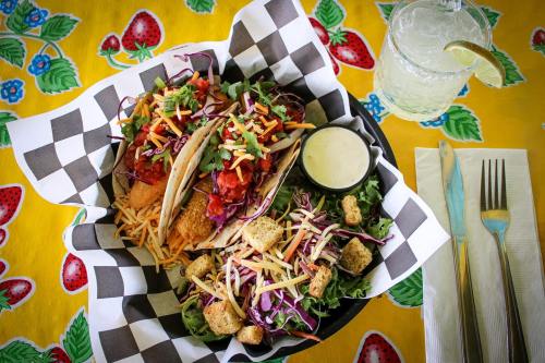 Two tacos filled with colorful toppings, served with a side of creamy sauce, on a checkered plate, with a drink nearby.