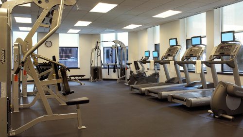 A modern gym with various exercise machines, including treadmills and weight equipment, under bright windows.