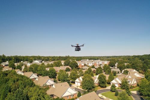 A drone hovers above a suburban neighborhood with houses and trees under a clear blue sky.