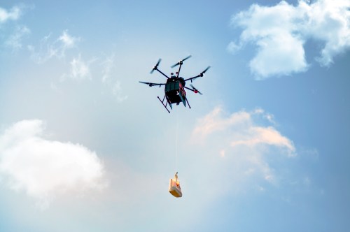 A drone flying in the sky, carrying a package suspended from a rope against a backdrop of clouds.
