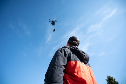 A person in a jacket looks up at a drone carrying a package against a clear blue sky.