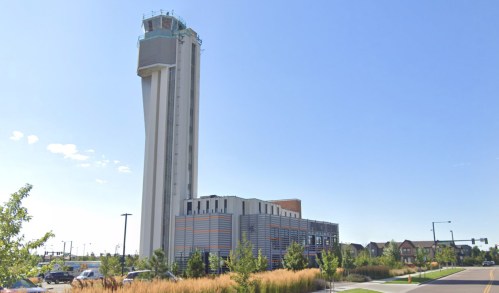 A tall airport control tower stands beside a modern building, surrounded by greenery and clear blue skies.