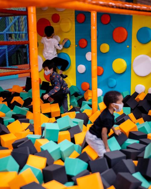 Children play in a colorful foam pit, climbing and exploring a wall with vibrant shapes while wearing masks.