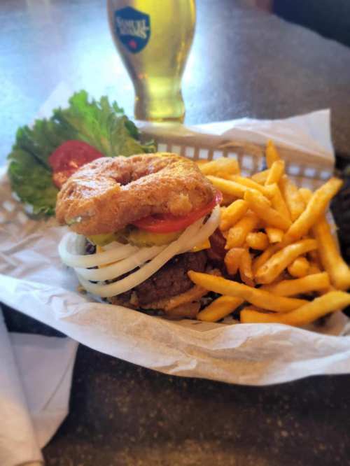 A burger with lettuce, tomato, onion, and pickles on a bagel, served with fries and a glass of beer in the background.