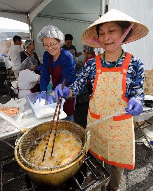 Two women cook at a food stall, one frying food in a large pot while wearing an apron and a traditional hat.