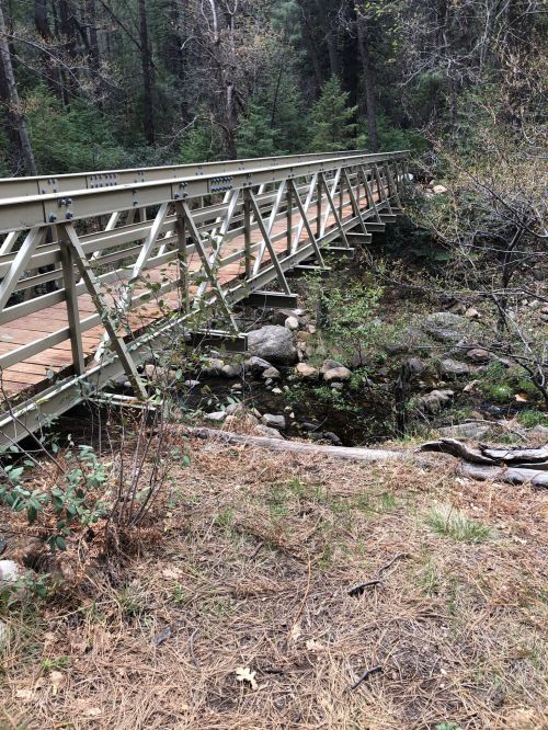 A metal bridge spans a small creek, surrounded by trees and rocky terrain in a forested area.