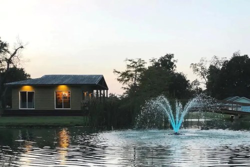 A serene lakeside scene featuring a house, a fountain with blue water, and trees at sunset.