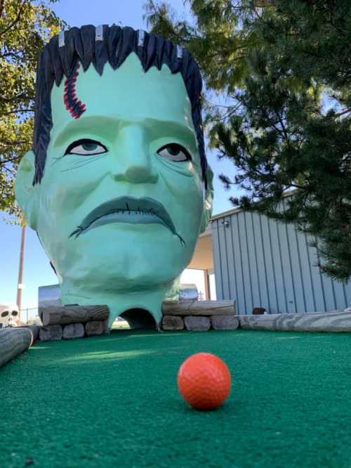 A giant Frankenstein head overlooks a mini-golf course, with a bright orange ball in the foreground on green turf.