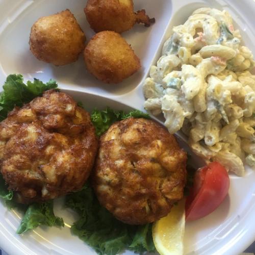 A plate featuring two crab cakes, macaroni salad, a slice of tomato, a lemon wedge, and three hushpuppies on lettuce.