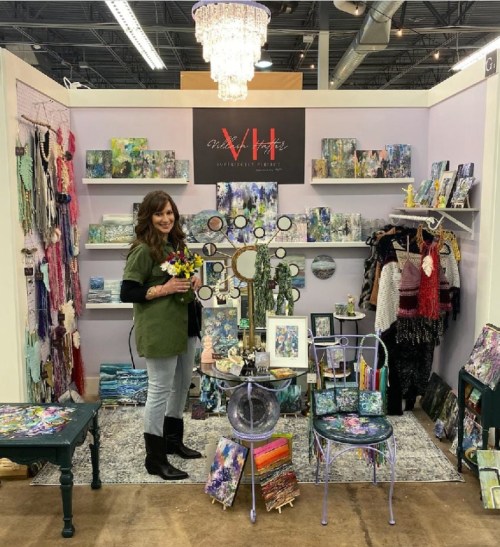 A woman stands in a colorful art booth filled with paintings, decor, and accessories, holding a bouquet of flowers.