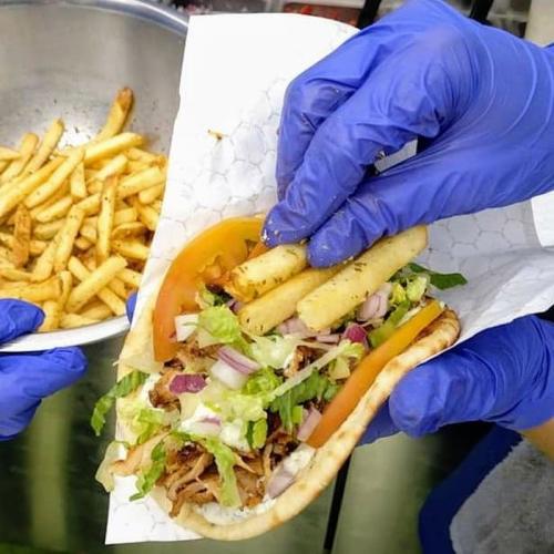 A close-up of hands in blue gloves assembling a sandwich with lettuce, tomatoes, and fries, with a bowl of fries in the background.