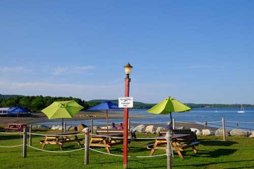 A scenic view of a beach with picnic tables and colorful umbrellas, overlooking calm water and distant sailboats.