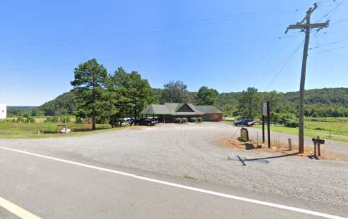 A gravel parking lot in front of a green-roofed building, surrounded by trees and hills under a clear blue sky.