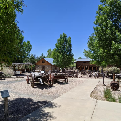 A sunny outdoor scene featuring historic buildings and vintage wagons surrounded by trees and a gravel path.