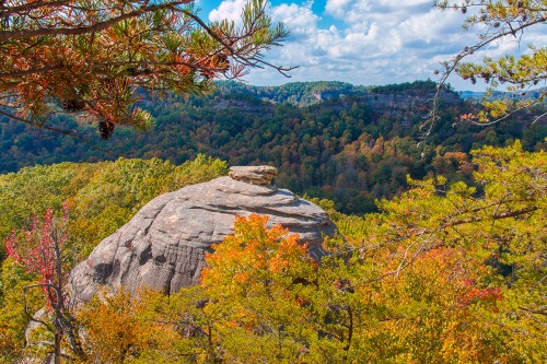 A rocky outcrop surrounded by vibrant autumn foliage and rolling hills under a blue sky with fluffy clouds.