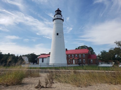 A tall white lighthouse with a red roof stands beside a sandy beach, surrounded by grass and trees under a blue sky.
