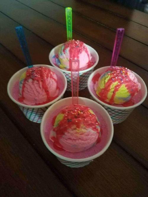 Four colorful ice cream cups with pink and yellow scoops, topped with sprinkles and syrup, on a wooden table.