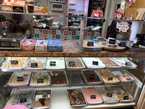 Display case filled with various types of fudge and candy, with labels and a scale in a cozy shop setting.