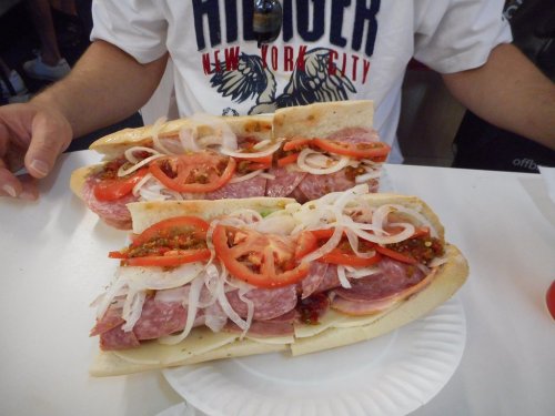 A person holding two large sandwiches filled with salami, cheese, tomatoes, onions, and peppers on a white plate.
