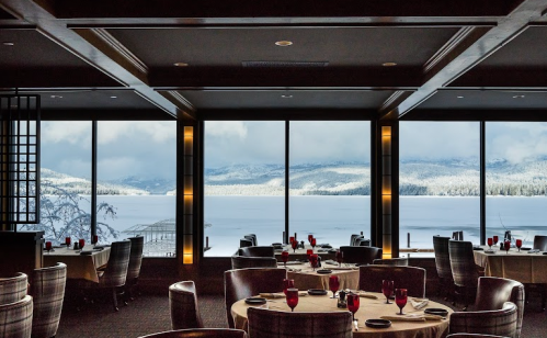 Elegant restaurant interior with round tables set for dining, overlooking a snowy lake and mountains through large windows.