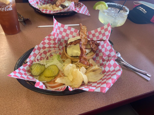 A burger with bacon and cheese, served with pickles, lettuce, tomato, and chips on a red-checkered plate.