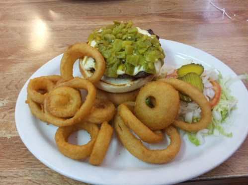 A plate with a cheeseburger topped with green peppers, served with onion rings and a side of lettuce, tomato, and pickles.
